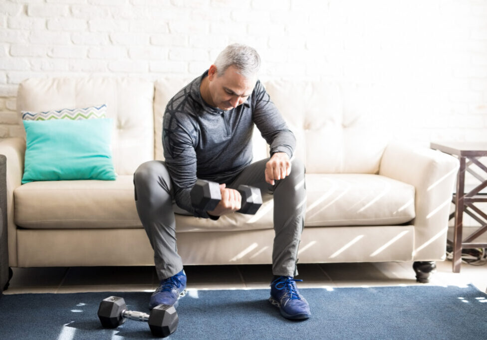 Hispanic man in his 50s sitting on sofa in living room and exercising, lifting dumbbells.